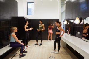 4 Reasons Why You Should Own a Gym Franchise 3 photo of five women in workout clothes talking in gym locker room with black tile walls.