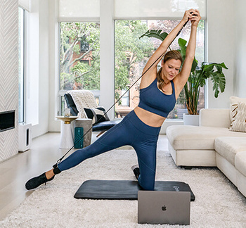 a woman in a blue sports top and leggings doing a leg exercise on a laptop