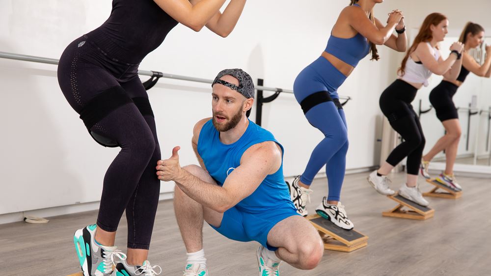 Four women working out and one male trainer checking their form and helping them.